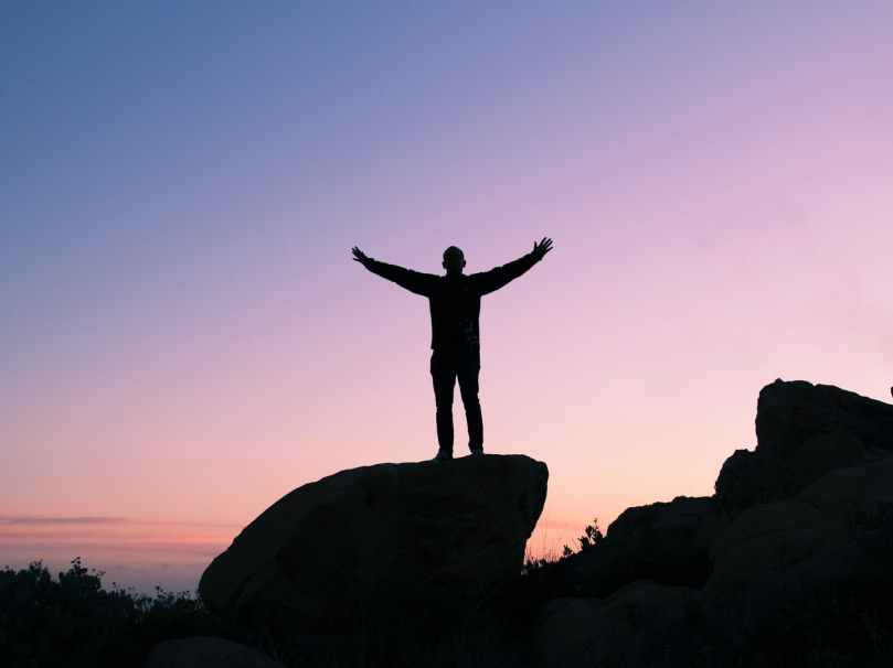 Person in shadow standing on rock with arms raised; the background is a blue and purple sky.